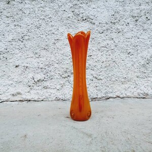 May include: An orange glass vase with a tall, slender body and a flared, petal-like top. The vase is approximately 12 inches tall and has a smooth, glossy finish. It stands on a concrete surface against a textured white wall.