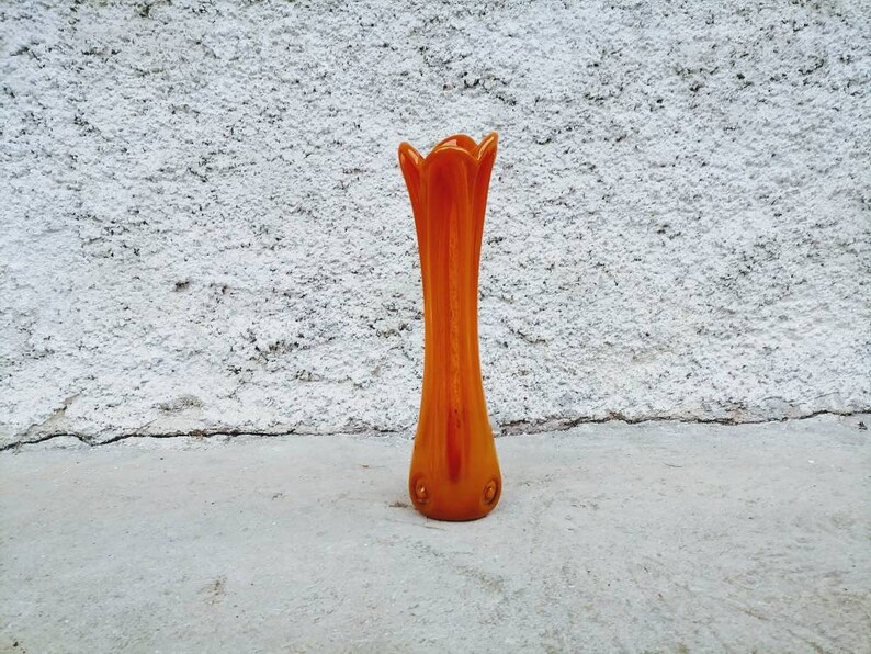 May include: An orange glass vase with a tall, slender body and a flared, petal-like top. The vase is approximately 12 inches tall and has a smooth, glossy finish. It stands on a concrete surface against a textured white wall.
