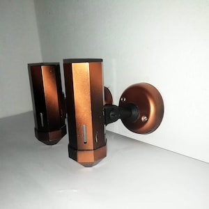 May include: Two copper-colored soap dispensers mounted on a white wall with a black metal bracket. The dispensers have a rectangular shape with a rounded top and bottom.