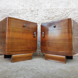 Könnte beinhalten: Zwei passende Nachttische aus Holz mit warmbraunem Finish. Jeder Schrank hat eine einzelne Tür mit einem kleinen Metallgriff und eine Schublade mit einem runden Knopf. Die Nachttische stehen auf einem rechteckigen Sockel.