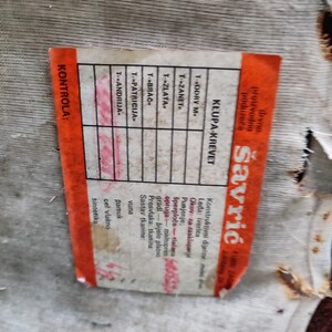 May include: A close-up of a red and orange label with the text "KLUPA-KREVET" and "Savrić" in white. The label also includes a table with the headings "T-ODRV M", "T-ZANET", "TZLATA", "T-BRAC", "T-PATRICIJA", "T-ANDRIJA" and a column with the headings "Konstruktivni dijelovi", "Leda", "Okov", "Punjenje", "Presvlaka", "Sastav tkanine".