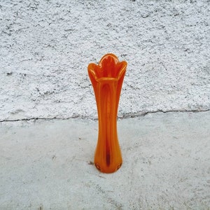 May include: An orange glass vase with a flared top and a slender, elongated body. The vase is approximately 12 inches tall and stands on a concrete surface against a textured white wall.