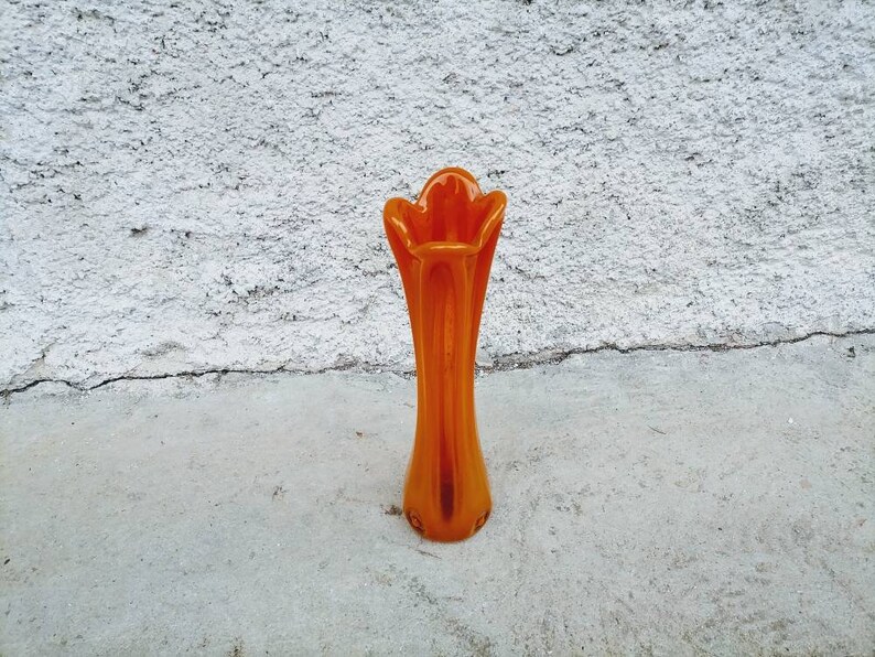 May include: An orange glass vase with a flared top and a slender, elongated body. The vase is approximately 12 inches tall and stands on a concrete surface against a textured white wall.