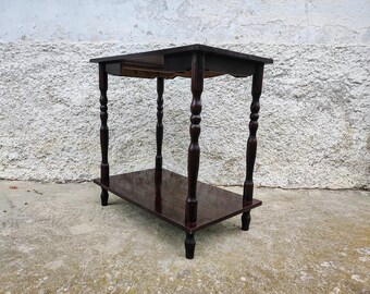 Puede incluir: Una mesa auxiliar de madera marrón oscuro con un estante y patas torneadas. La mesa tiene un diseño sencillo y está hecha de madera maciza.