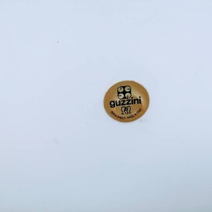 Może przedstawiać: Złota naklejka z tekstem "Guzzini" i słowami "Forniture Made in Italy" w k&oacute;łku.