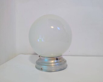 May include: A vintage-style, frosted glass globe light fixture. The spherical glass shade sits atop a circular, silver-toned metal base. The light fixture is designed for indoor use and provides soft, diffused lighting.