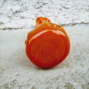 May include: A small, orange, ceramic sculpture with a glossy finish. The rounded form has a smooth surface with a small, raised detail on top. The sculpture is set against a textured, light gray background.