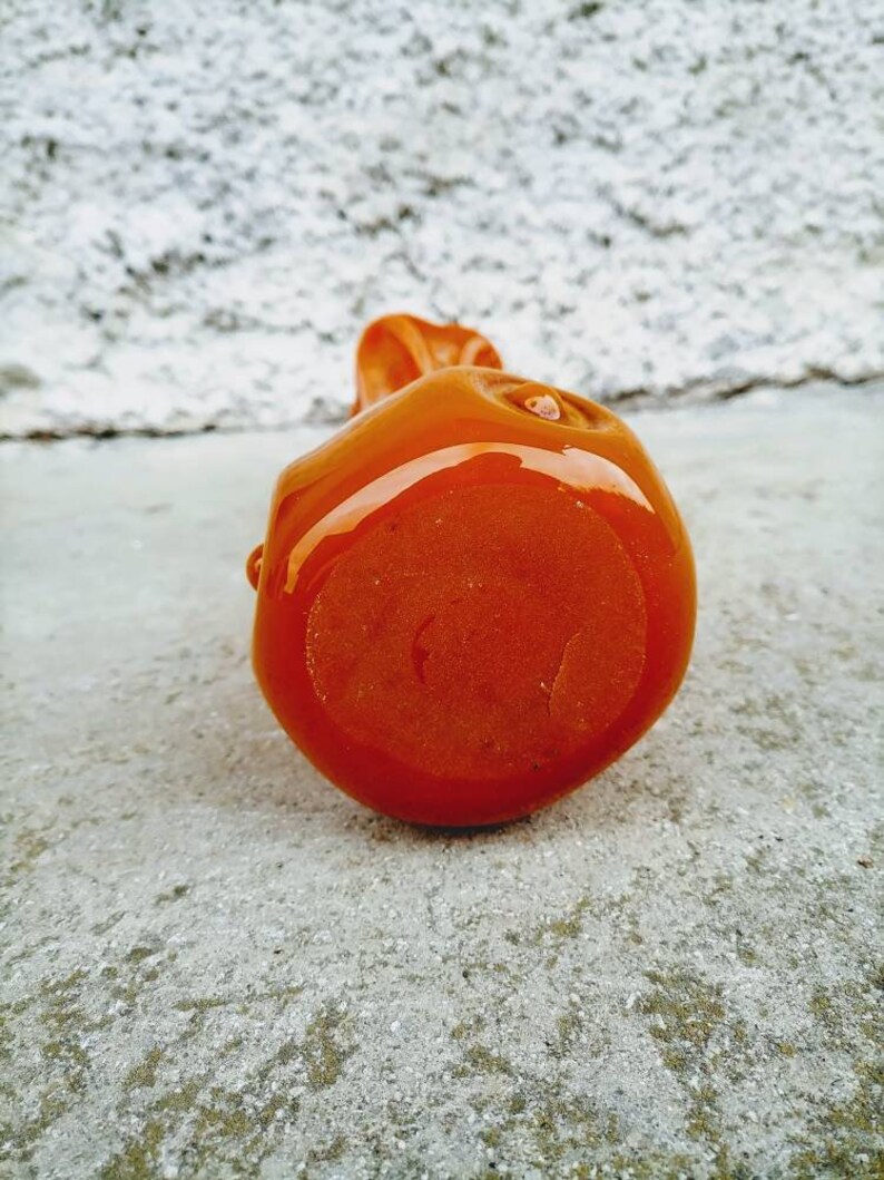 May include: A small, orange, ceramic sculpture with a glossy finish. The rounded form has a smooth surface with a small, raised detail on top. The sculpture is set against a textured, light gray background.