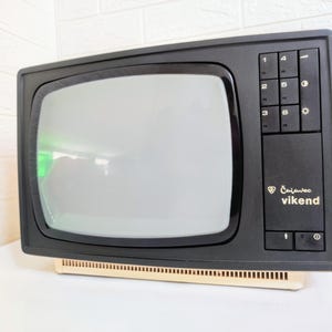 以下が含まれることがあります： ヴィンテージの黒とクリーム色のテレビ。画面は明るいグレーです。テレビの右側には一連のボタンがあり、前面には「Čajovec vikend」の文字が印刷されています。