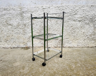 May include: A chrome and glass rolling cart with three shelves. The cart has four black wheels and is standing on a concrete surface.