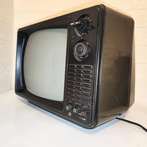Puede incluir: Un televisor negro vintage con una pantalla redondeada y perillas de control. El televisor tiene un marco oscuro y una serie de botones y puertos en la parte delantera. La pantalla es de color gris claro. El televisor está sobre una superficie blanca.