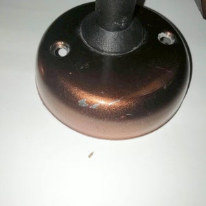 May include: A copper-colored metal base with a black plastic post extending from the center. The base has two small holes on the side and a small inscription on the bottom.