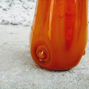 May include: A close-up of a vibrant orange glass vase with a glossy finish. The vase has a rounded shape with a flared top and a unique swirl design at the base. The glass reflects light, highlighting its smooth texture and color.