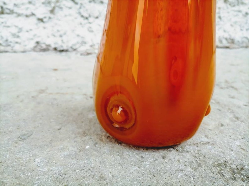 May include: A close-up of a vibrant orange glass vase with a glossy finish. The vase has a rounded shape with a flared top and a unique swirl design at the base. The glass reflects light, highlighting its smooth texture and color.