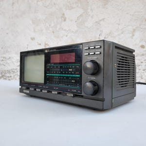 以下が含まれることがあります： ヴィンテージのグレーのポータブルテレビとラジオのコンボユニット。このデバイスは、小さな画面、ラジオダイヤル、およびさまざまなコントロールノブを備えています。このユニットには、スピーカーと電源コードが内蔵されています。