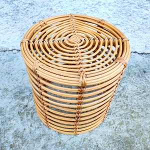 May include: A cylindrical, woven rattan side table. The natural-toned table features a circular top and is constructed with horizontal and vertical rattan strips. The design is a light brown color and is suitable for indoor or outdoor use.