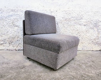 Könnte beinhalten: Ein grau gepolsterter Sessel mit hohem Rücken und kantigem Design. Der Stuhl hat einen strukturierten Stoff und steht vor einer strukturierten weißen Wand. Der Stuhl ist ein Einsitzer.