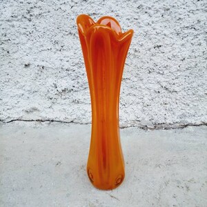 May include: An orange glass vase with a flared top and a slender body. The vase has a smooth, glossy finish and is set against a textured white background. The vase is approximately 12 inches tall.