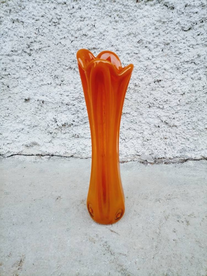 May include: An orange glass vase with a flared top and a slender body. The vase has a smooth, glossy finish and is set against a textured white background. The vase is approximately 12 inches tall.