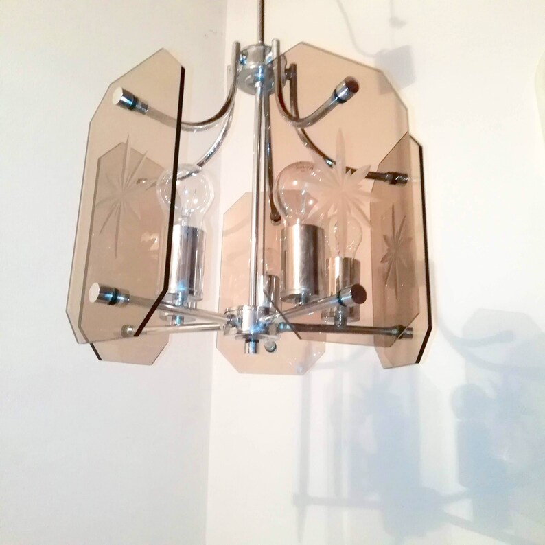 May include: A vintage chandelier with a chrome frame and amber-colored glass panels. The panels are etched with starburst designs. The light fixture has three exposed light bulbs.