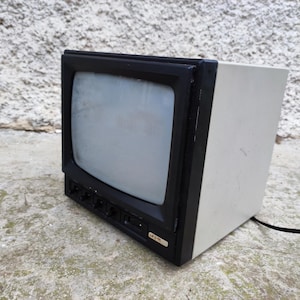 以下が含まれることがあります： 黒いフレームと明るい色のボディを持つ、古い箱型のテレビ。画面は明るいグレーです。テレビは粗いコンクリートの表面に置かれています。黒い電源コードが背面から伸びています。