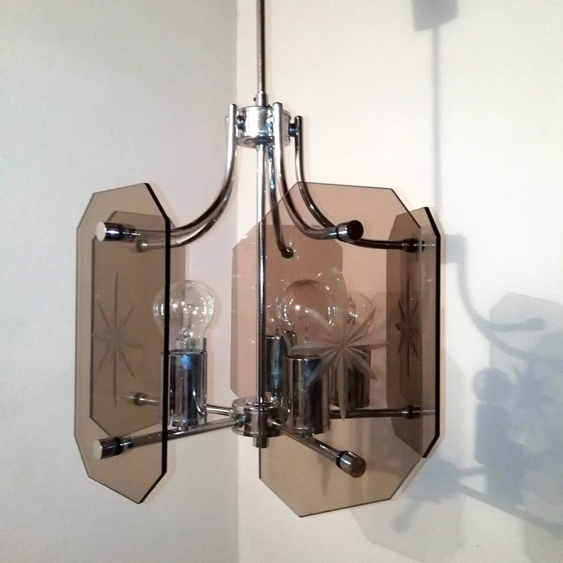 May include: A vintage chrome and glass chandelier with a mid-century modern design. The fixture features three light bulbs, etched starburst designs, and geometric, smoky-brown glass panels. The light fixture is suspended from a ceiling.