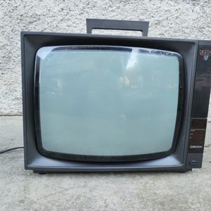 Puede incluir: Un televisor Orion gris oscuro vintage con un asa de transporte prominente en la parte superior. El frente presenta una gran pantalla de cristal, un panel de control negro en el lado derecho con el texto DIGITAL y ORION, y un botón de encendido.