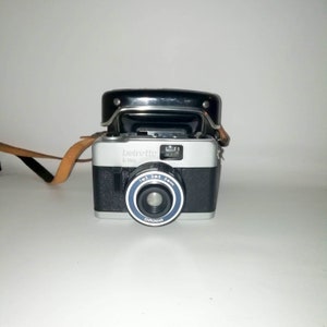 以下が含まれることがあります： 黒い革のストラップが付いた銀と黒の35mmフィルムカメラ。カメラには青いリングが付いた黒いレンズがあり、レンズには「Chromar」という文字が印刷されています。カメラには「beirette K 200」という文字も印刷されています。