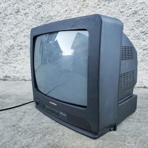 以下が含まれることがあります： 黒いGoldStar製のブラウン管テレビ。かさばるプラスチック製の筐体、湾曲したガラススクリーン、ボタンと電源インジケーターライトを備えた前面コントロールパネルが特徴で、背面からは細い黒い電源コードが伸びています。