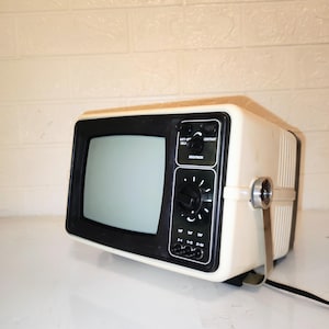 Könnte beinhalten: Vintage tragbarer Fernseher in cremefarbener Ausführung mit schwarzem Bildschirm und Bedienfeld. Der Fernseher hat eine Holzoptik-Oberseite und einen silbernen Griff. Das Bedienfeld enthält Knöpfe für Helligkeit und Kontrast. Ein schwarzes Netzkabel.