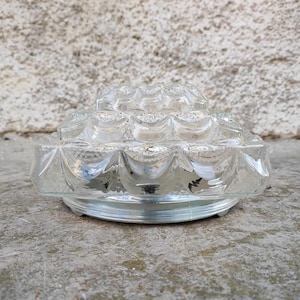 May include: A clear, glass light fixture with a textured, geometric pattern. The fixture has a tiered design and sits on a silver-colored base. The glass has a reflective quality, and the fixture is likely for a ceiling or wall.