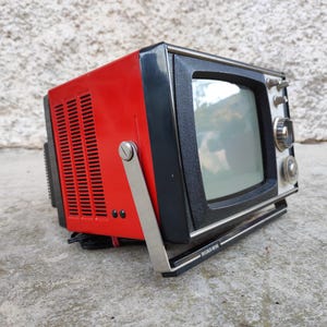 Könnte beinhalten: Ein tragbarer Vintage-Fernseher mit leuchtend rotem Gehäuse und schwarzer Frontblende. Das Gerät verfügt über einen silbernen Metalltragegriff, einen kleinen Bildschirm und runde Drehknöpfe auf der rechten Seite sowie einen schwarzen Sockel.