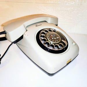Puede incluir: Un teléfono de disco rotatorio vintage blanco roto con un dial negro y un cable en espiral negro. El teléfono tiene una base rectangular y un auricular curvo. El dial presenta números y un dial central. Un clásico de la tecnología de comunicación.