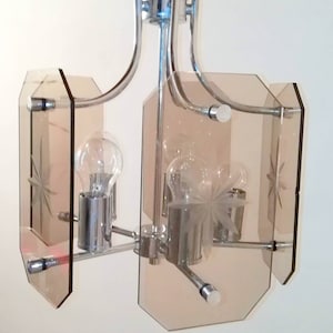 May include: A chrome and glass chandelier with a mid-century modern design. The fixture features four light bulbs and four amber-colored glass panels with etched starburst patterns. The metal frame has curved arms.