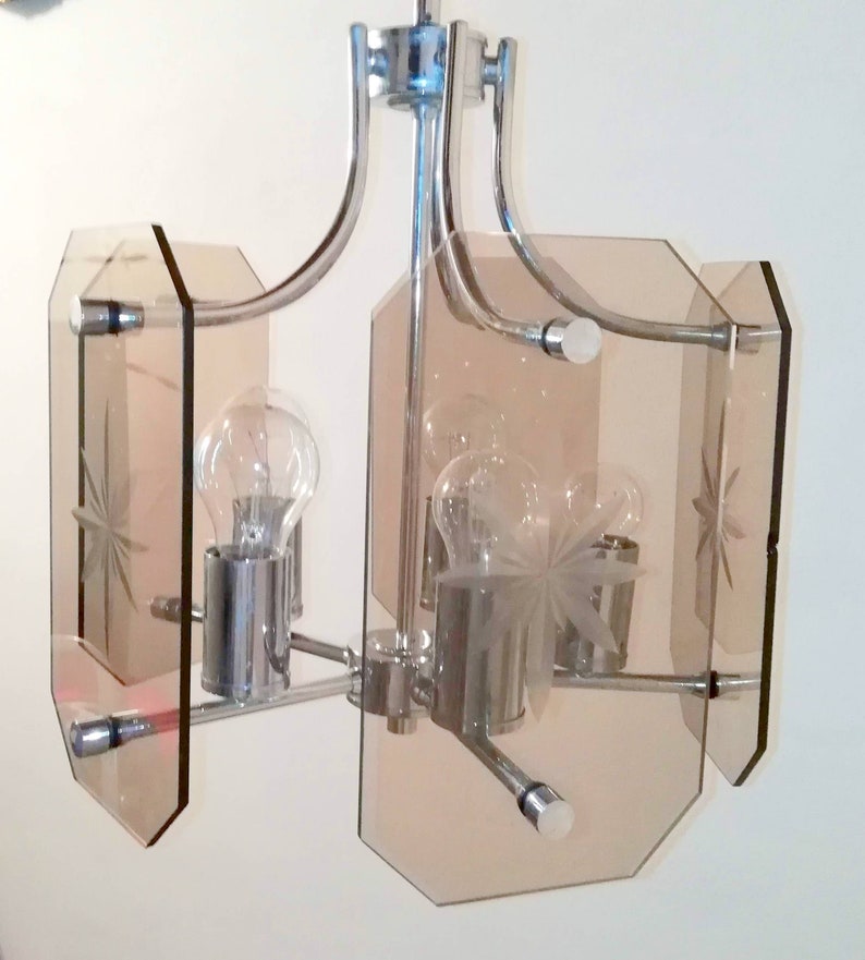 May include: A chrome and glass chandelier with a mid-century modern design. The fixture features four light bulbs and four amber-colored glass panels with etched starburst patterns. The metal frame has curved arms.