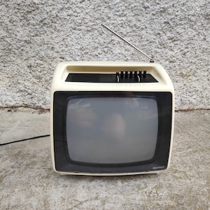 May include: A vintage portable television with a cream-colored plastic casing and a black screen frame. The device features a top-mounted extendable metal antenna, a series of black control knobs, and a black power cord extending from the side.