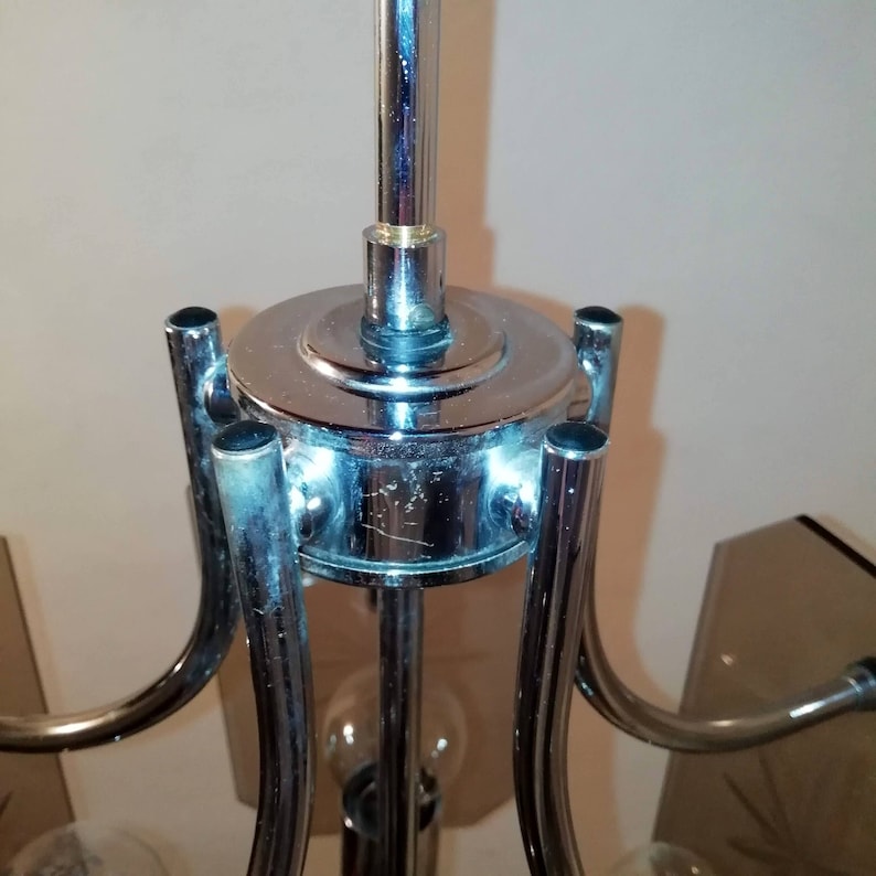May include: Close-up of a chrome chandelier fixture. The fixture has a central vertical pole with a circular base and curved arms extending outwards. The metal has a reflective surface, and the design is modern.