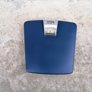 Könnte beinhalten: Eine blaue Badezimmerwaage mit silbernem Zifferblatt und verchromtem Rand. Die Waage hat ein Zifferblatt mit Zahlen von 0 bis 250.