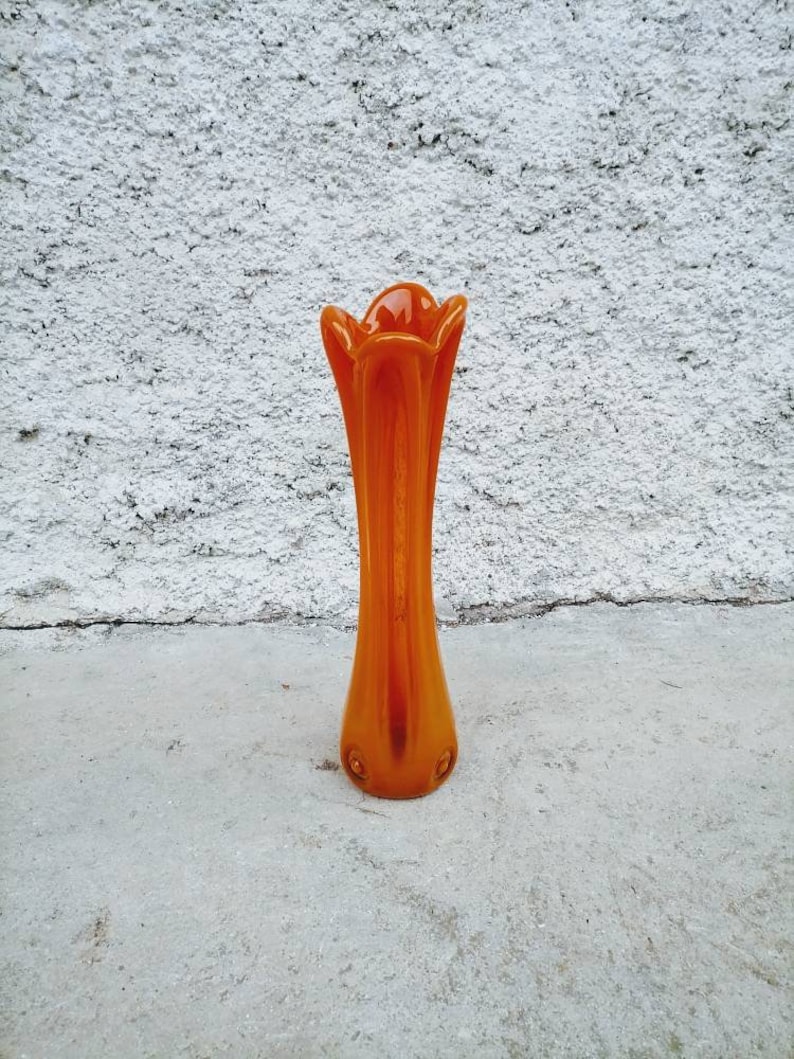 May include: An orange glass vase with a flared top and a slender body. The vase has a glossy finish and is set against a textured white wall. The vase is approximately 10 inches tall.