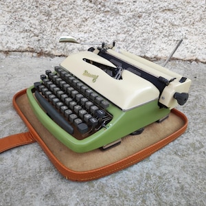 May include: Vintage typewriter in a cream and green color scheme, resting on a brown leather case. The typewriter features a black keyboard and a chrome paper holder. The brand name is visible on the top.
