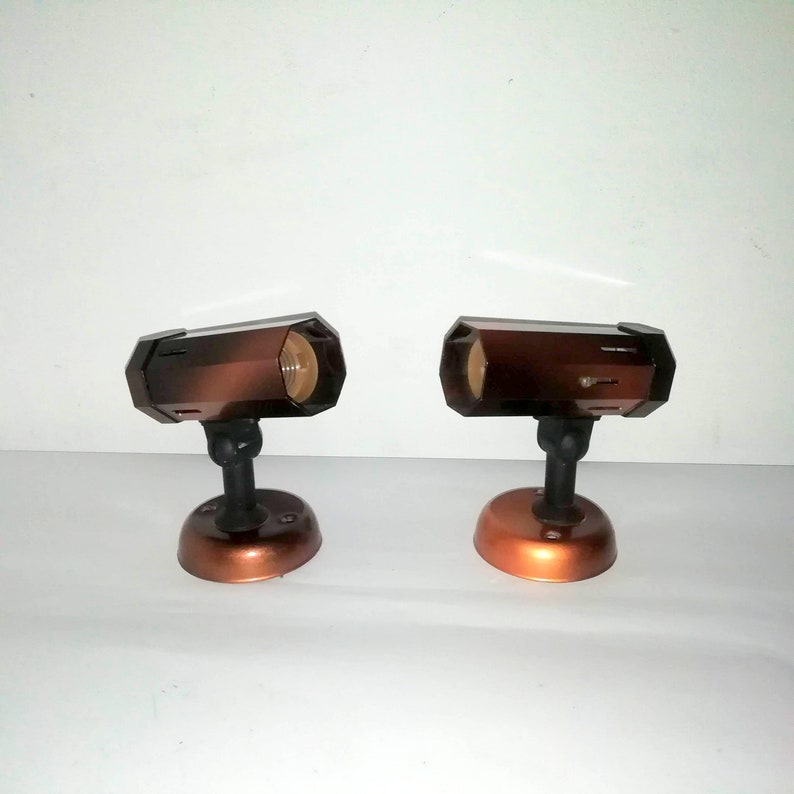 May include: Two brown metal spotlights with a round base and a swiveling head. The spotlights are on a white background.