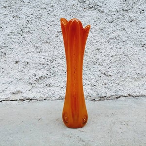 May include: An orange glass vase with a tall, slender shape and a flared top. The vase has a smooth, glossy finish and is approximately 12 inches tall. The base features two small, circular decorative elements.