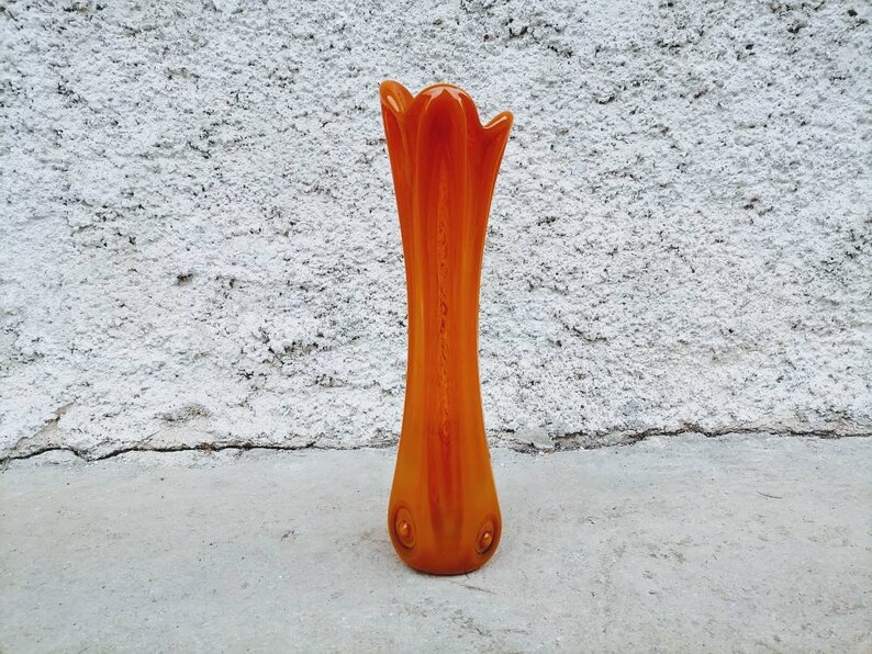 May include: An orange glass vase with a tall, slender shape and a flared top. The vase has a smooth, glossy finish and is approximately 12 inches tall. The base features two small, circular decorative elements.