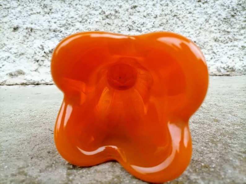 May include: An orange, glossy, flower-shaped object. The object has a smooth, reflective surface and a rounded, organic form. The interior is visible, revealing a hollow space. The object is set against a textured, light-colored background.