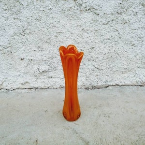 May include: An orange glass vase with a flared, petal-like top. The vase has a tall, slender shape and is made of translucent glass. The vase is approximately 10 inches tall and is set against a textured white wall.