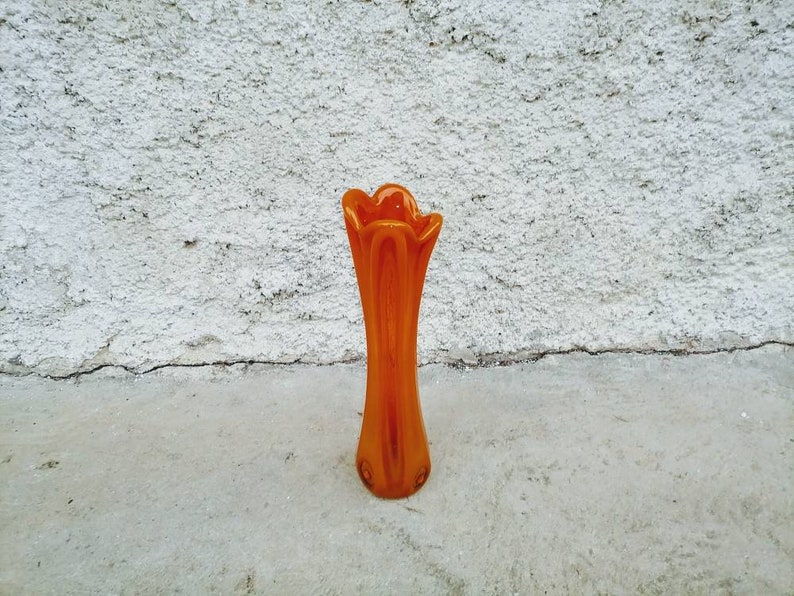 May include: An orange glass vase with a flared, petal-like top. The vase has a tall, slender shape and is made of translucent glass. The vase is approximately 10 inches tall and is set against a textured white wall.