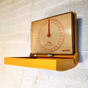 Op de afbeelding: Vintage gele en crèmekleurige keukenweegschaal bevestigd aan een witte bakstenen muur. De wijzerplaat heeft een ronde schaal met rode markeringen en een rode naald, die tot 1000 gram meet. De weegschaal heeft een gele bak en de merknaam "gorenje".