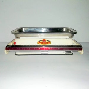 Pode incluir: Balança de cozinha branca com uma bandeja de aço inoxidável e uma escala vermelha e preta. A balança tem a palavra "Prima" em letras vermelhas sobre um círculo amarelo.