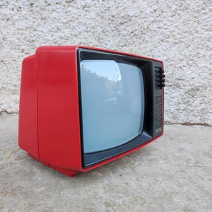 May include: A vintage portable television with a bright red plastic casing and a black front panel surrounding the screen. The device features a small speaker grille and a vertical row of black control knobs on the right side of the display area.
