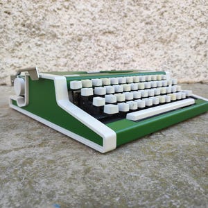 May include: A vintage green typewriter with white keys and trim. The typewriter is set on a concrete surface. The keys are arranged in a standard QWERTY layout. The machine has a classic, retro design.